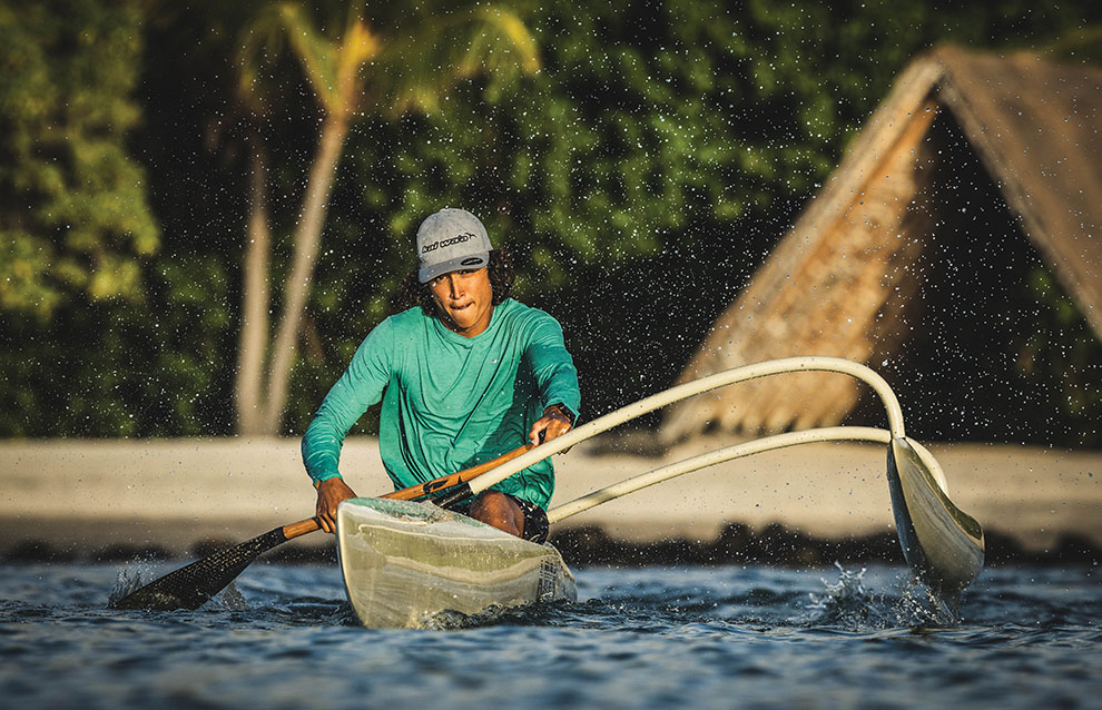person in teal shirt and baseball cap canoes away from land. 